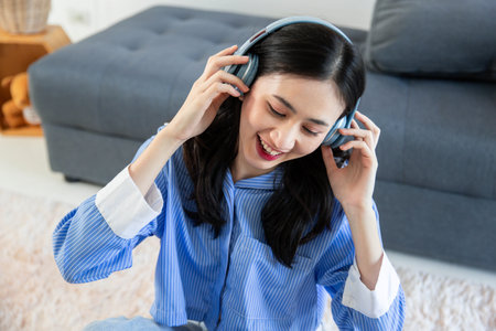 Beautiful asian woman listening to music on couch in living room at home. Happy asia female wearing headset and sitting on sofaの写真素材