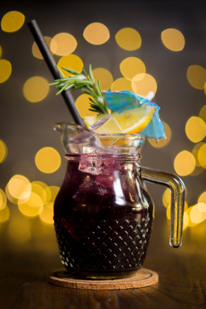 Cold refreshing, grape juice in jug with slice of lemon, sprig of rosemary and small umbrella decoration, on blurred lights backgroundの写真素材