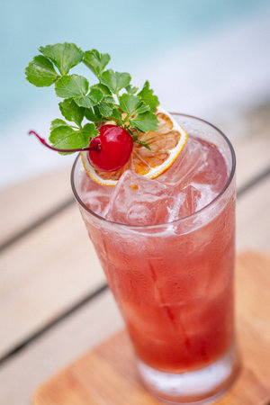 High angle shot, Sex on the beach cocktail in glass garnished with a slice of lemon, a cherry, and a sprig of mint on wooden planks in front of a poolの写真素材