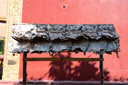 Wooden structure holding a layered arrangement of large and brown leaves for a roofing, background features a vibrant red wallの写真素材