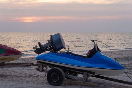 Blue jet ski on a trailer parked on a sandy beach with ocean and sunset sky in background at Chonburi, Thailandの写真素材