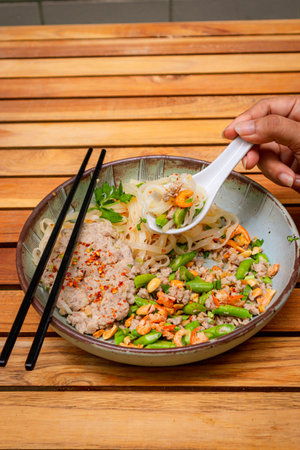 Noodles mince pork with chili and thai herbs in bowl placed on a wooden table, hand is holding a spoon and taking some noodles from the bowl.の写真素材