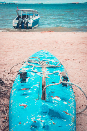 Close-up view of weathered blue surfboard resting on a sandy beach with a calm body of water in the...