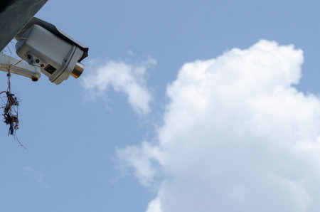 Low-angle, close-up view of a CCTV on a pole against a fluffy cloud and blue sky.