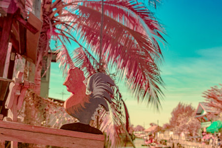 Infrared photography, a wooden rooster figurea, colorful background with palm trees and a body of waterの写真素材