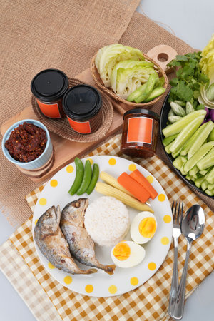 A meal setup featuring a cup of thai chili paste, a plate with two pieces of cooked fish, white rice, boiled egg and vegetables on wooden trayの写真素材