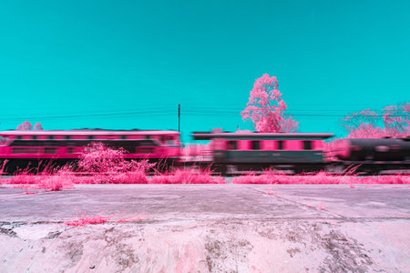 Infrared photography, trains in motion, captured in a vibrant color scheme with dominant shades of turquoise and pink. The background includes trees and power linesの写真素材