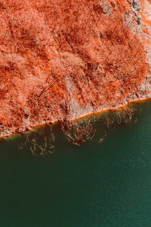 Infrared style, a landscape with a steep, reddish-brown terrain on one side and a calm body of water on the otherの写真素材