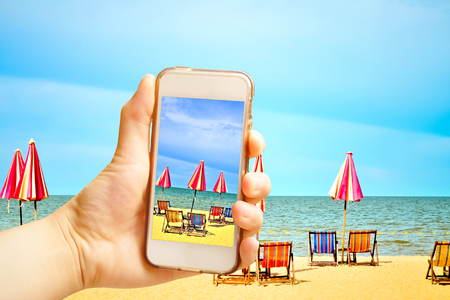 Hand holding smart phone (Mobile Phone) with Chairs and umbellar on white sand beach.の写真素材