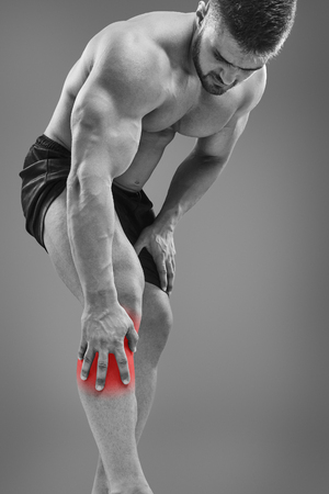 Fit attractive man having a knee pain, bending over the leg, isolated on gray background. Highlighted red spot conceptの写真素材