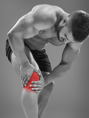 Bodybuilder gripping an inflamed knee, isolated on gray background. Highlighted red spot conceptの写真素材
