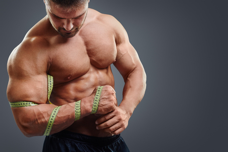Closeup of bodybuilder holding tape measure. Wrong biceps measuring concept. Muscular young man tries to measure his muscle bicep by himself isolated over gray background. Wrong muscle measuringの写真素材