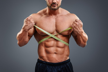 Personal fitness trainer holding tape measure isolated over gray background. Closeup of muscular man measuring his waist. Doing perfect six pack abs.の写真素材