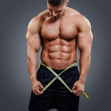 Fitness male model measuring his waist with tape measure isolated over gray background. Gain in abdominal muscles concept.の写真素材