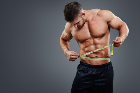 Muscular sports man measuring his waist with tape measure isolated over gray background. Bodybuilder waist measure concept.の写真素材