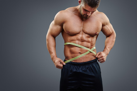 Fitness male model measuring his waist with tape measure isolated over gray background. Gain in abdominal muscles concept.の写真素材