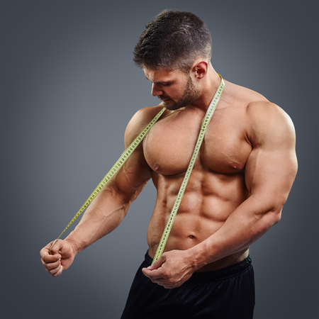 Muscular sports man tries to measure his neck muscles with measuring tape isolated over gray background. Confused bodybuilder holding tape measure.の写真素材