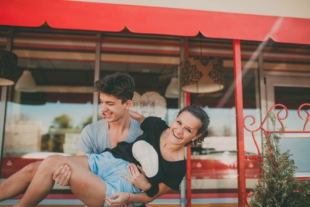 Couple in love having fun outside a cafe. Handsome young man holding his girlfriend on hands. Young teenage people laughingの写真素材