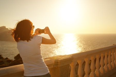 Young woman taking picture of sunset at Cap Formentor, Majorca. Girl photographing sunset with her phoneの写真素材