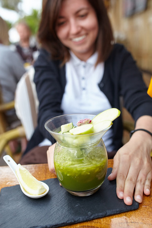 Smiling young woman eating green gazpacho soup at restaurant. Fresh raw vegetarian cold soup.の写真素材