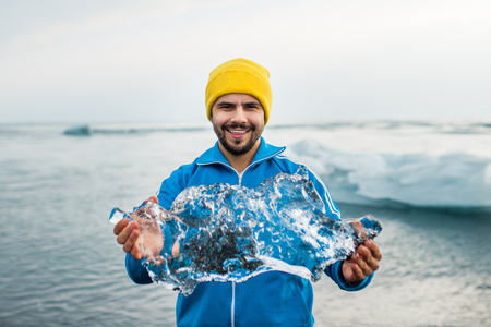 Closeup of smiling young man holding a piece of ice on black sand beach. Male holding piece of iceberg. Iceland vacation. Travel to Iceland.の写真素材