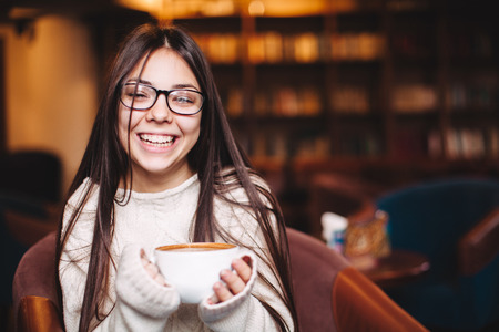 Pretty smiling girl wearing eyeglasses sitting and holding a cup of coffee with copyspace. Happy teenage girl laughing with mug of coffeeの写真素材