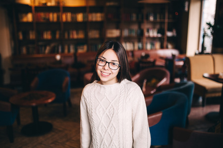 beautiful asian young woman posing inside a cafe. Teenage girl wearing eyeglasses and sweater standing indoors.の写真素材