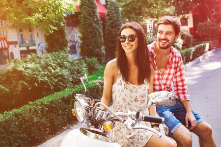 glamorous  young couple sitting on a vintage scooter in the street, man wears a shirt and the woman - a summer dress and sunglassesの写真素材