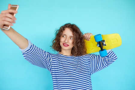 Vivid portrait of hipster pretty cool young woman with skateboard taking picture self portrait on smartphone isolated over blue backgroundの写真素材