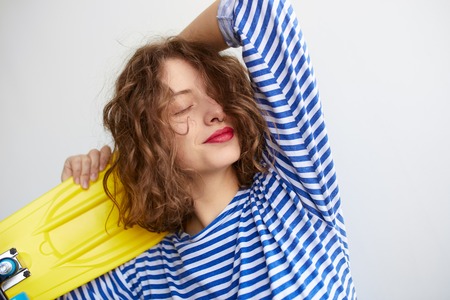 Closeup of beautiful girl with curly hairstyle and red lips holding a yellow skateboard on white background. Hipster girl dreaming with eyes closed.の写真素材