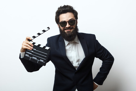 Smiling man with beard and sunglasses wearing an elegant black suit holding holding film clapper over white background. Film director concept.の写真素材
