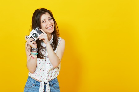 Summer portrait of stylish pretty hipster girl with retro vintage camera posing over yellow background with copy space.の写真素材