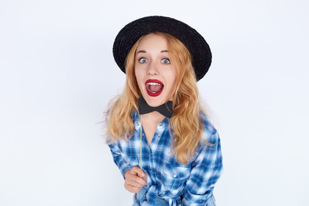 Stylish fashion portrait of trendy casual young woman in blue shirt and black hat holding a paper bow tie, Hipster blonde girl posing with surprised face isolated on white background.の写真素材