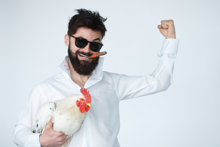 crazy young man showing bicep muscle and holding a cock chicken on white background. Cuban bearded man wearing sunglasses and smoking cigarの写真素材
