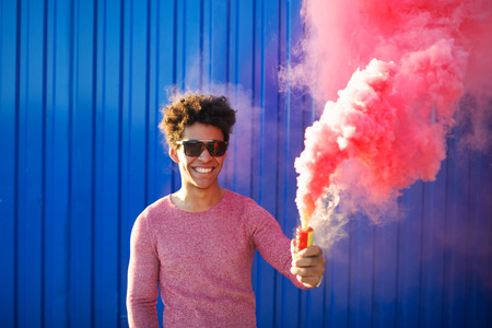 Portrait of a young black hipster man smiling and holding a colorful pink smoke bomb over a blue color background. Youth and lifestyle conceptの写真素材