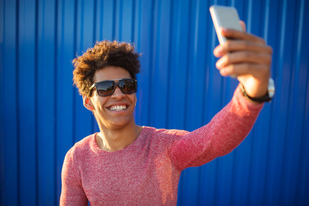 young happy man casual dressed with smart phone on vivid blue background. Colorful concept with african american teenage man taking a selfieの写真素材