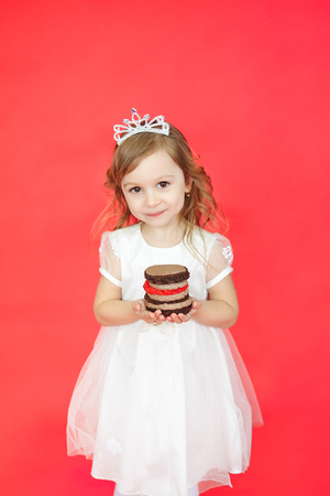Vertical portrait of blond hair little girl holding a gift cake on red background. Cute preschoolder wearing a princess crown and white beautiful dress have a presentの写真素材