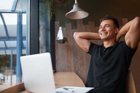 Satisfied with work done. Happy smiling young man working on laptop keeping hands behind his head. Being productive at cafe. Copy spaceの写真素材