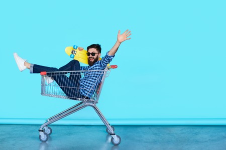 Carefree hipster fun. Side view of cheerful young man sitting in shopping cart with skateboard over blue wall with copy space.の写真素材