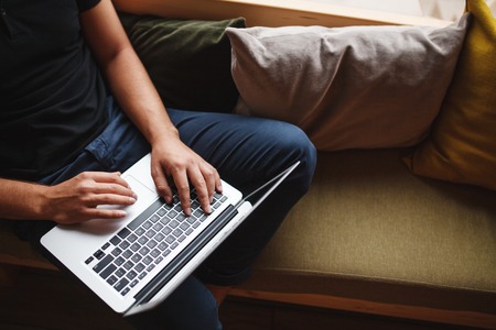 Portrait from above man sitting on sofa with laptop with copy space. Close up male hands typing on portable computer keyboardの写真素材