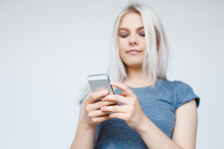 Selective focus on smartphone. Casual young woman with blonde hair texting on her phone - over white background with copyspace. Low angle viewの写真素材