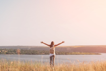 Back view of beautiful girl in the meadow with travel backpack holding hands raised. Freedom conceptの写真素材