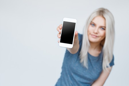 Teenage girl showing a blank smartphone screen. Focus on smartphone. Cute young blond student in backgroundの写真素材