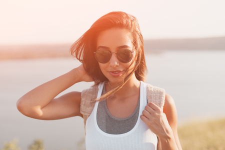 Summer sunny lifestyle fashion portrait of young stylish hipster woman wearing sunglasses, travel with backpack. Sun flare effect. Toned imageの写真素材