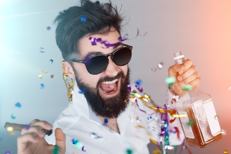Happy smiling man celebrating and partying with smoking cigar and whiskey bottle in his hand with light flare effectの写真素材