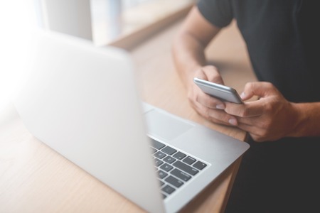 man's hands using smart phone at laptop in interior, young male student typing on phone sitting at...