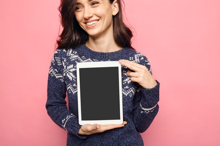 Woman holding a tablet with empty black screen over pink color background. Smiling girl in winter clothes showing display with copy space for app contentの写真素材