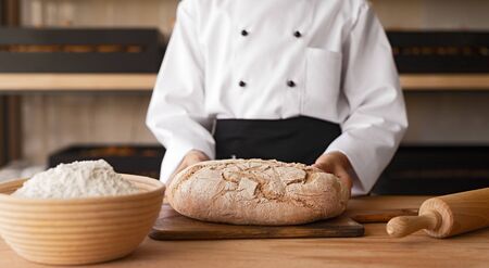 Unrecognizable kid in chef uniform putting loaf of bread on table while working in kitchen of cozy...