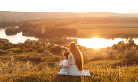 Back view of young girl embracing little brother while having happy moments together sitting on hill looking at sunset in calm river, flowing among green fields and forestの写真素材