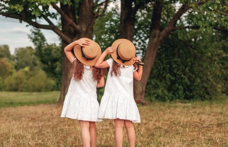 Back view of unrecognizable sisters in similar dresses and hats admiring tall tree on summer day in green parkの写真素材